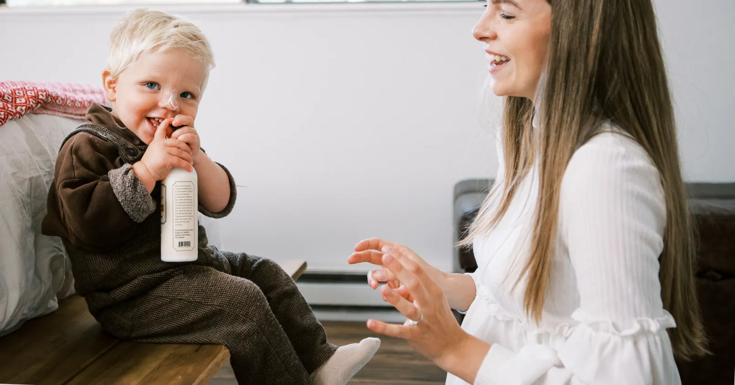 A young boy tries to taste goat milk lotion as mother laughs